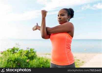 fitness, sport and healthy lifestyle concept - young african american woman stretching arms on beach. young african american woman stretching on beach