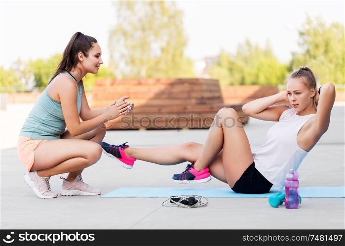 fitness, sport and healthy lifestyle concept - young woman assisting her friend doing bicycle crunches on mat outdoors. women training and doing bicycle crunches outdoors