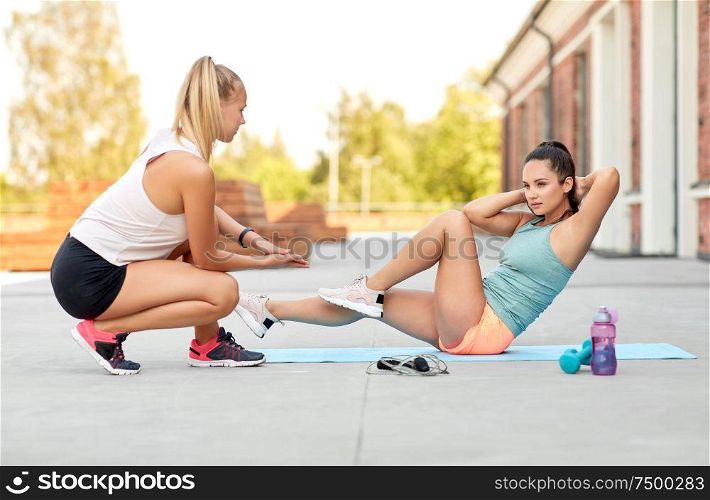 fitness, sport and healthy lifestyle concept - young woman assisting her friend doing bicycle crunches on mat outdoors. women training and doing bicycle crunches outdoors