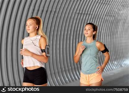 fitness, sport and healthy lifestyle concept - young women or female friends with earphones wearing armbands with smartphones and running outdoors. young women with earphones and smartphones running