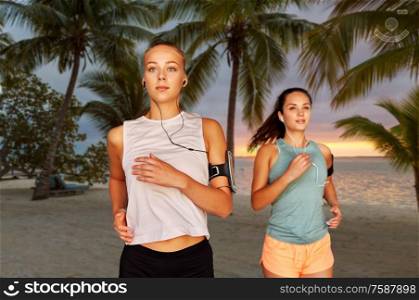 fitness, sport and healthy lifestyle concept - young women or female friends with earphones wearing armbands with smartphones and running over palm trees on tropical beach on background. young women with earphones and smartphones running