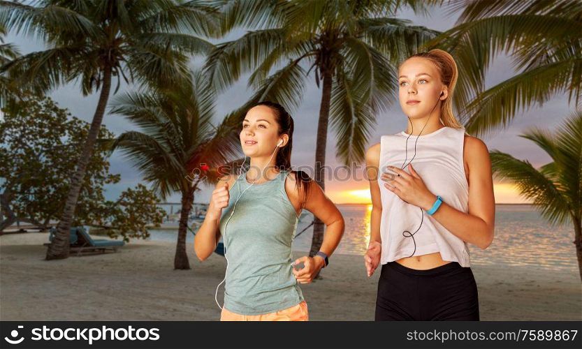 fitness, sport and healthy lifestyle concept - young women or female friends with earphones running over palm trees on tropical beach on background. women or female friends with earphones running