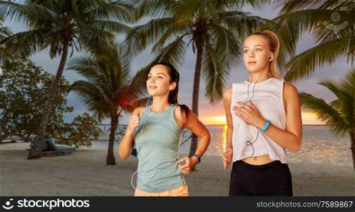 fitness, sport and healthy lifestyle concept - young women or female friends with earphones running over palm trees on tropical beach on background. women or female friends with earphones running