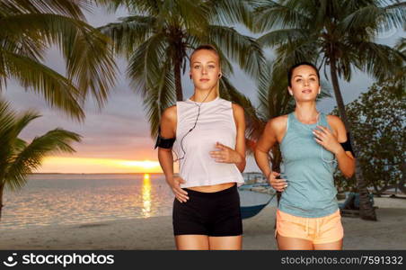 fitness, sport and healthy lifestyle concept - young women or female friends with earphones wearing armbands with smartphones and running over palm trees on tropical beach on background. young women with earphones and smartphones running