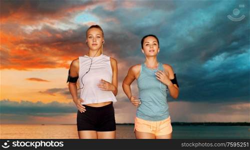 fitness, sport and healthy lifestyle concept - young women or female friends with earphones wearing armbands with smartphones and running over sea and sunset sky on background. young women with earphones and smartphones running