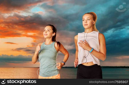 fitness, sport and healthy lifestyle concept - young women or female friends with earphones running over sea and sunset sky on background. women or female friends with earphones running