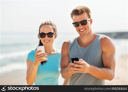 fitness, sport and lifestyle concept - happy couple in sports clothes and sunglasses with smartphones on beach. couple in sports clothes with smartphones on beach