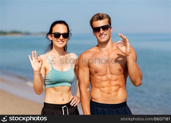 fitness, sport and lifestyle concept - happy couple in sports clothes and sunglasses on beach showing ok hand sign. couple of athletes in shades on beach showing ok