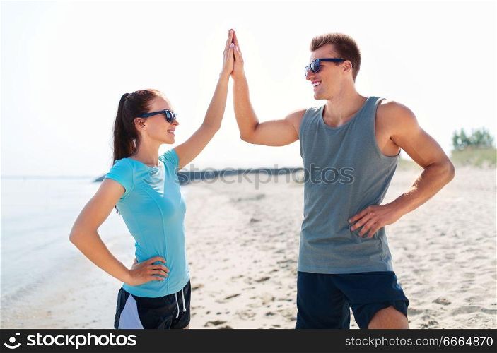 fitness, sport and lifestyle concept - happy couple in sports clothes and sunglasses on beach making high five gesture. happy couple in sports clothes and shades on beach