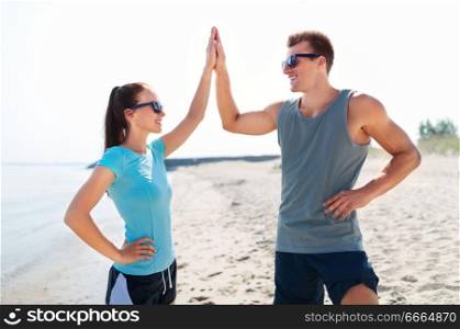 fitness, sport and lifestyle concept - happy couple in sports clothes and sunglasses on beach making high five gesture. happy couple in sports clothes and shades on beach