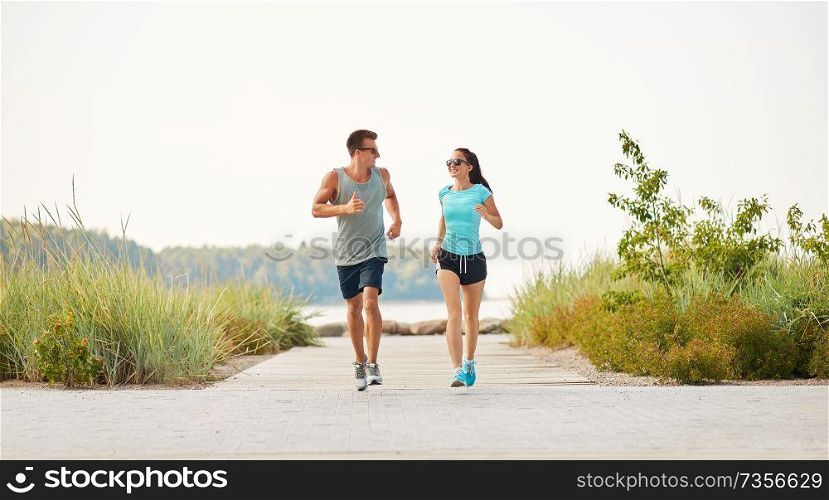 fitness, sport and lifestyle concept - happy couple in sports clothes and sunglasses running along summer beach path. couple in sports clothes running along beach path