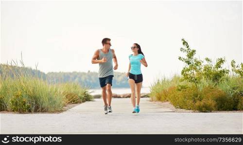fitness, sport and lifestyle concept - happy couple in sports clothes and sunglasses running along summer beach path. couple in sports clothes running along beach path