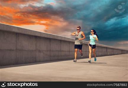 fitness, sport and lifestyle concept - happy couple in sports clothes and sunglasses running along concrete pier over sunset sky on background. couple in sports clothes running along pier