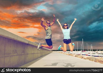 fitness, sport and lifestyle concept - happy couple in sports clothes jumping on pier over harbor and sunset sky on background. happy couple in sports clothes jumping on pier