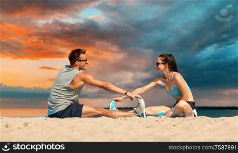 fitness, sport and lifestyle concept - smiling couple stretching legs on beach before training over sea and sunset sky on background. smiling couple stretching legs on beach