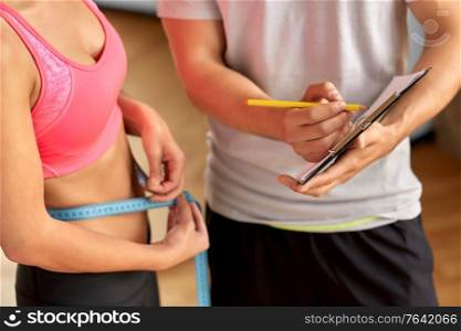 fitness, sport and people concept - close up of young woman with measuring tape and personal trainer with clipboard in gym. close up of woman with personal trainer in gym