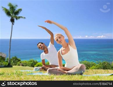 fitness, sport and people concept - couple making yoga exercises outdoors over exotic natural background with palm tree and ocean. happy couple making yoga exercises outdoors