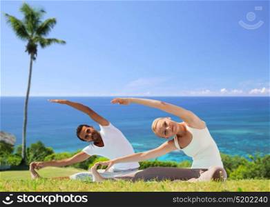 fitness, sport and people concept - couple making yoga exercises outdoors over exotic natural background with palm tree and ocean. happy couple making yoga exercises outdoors