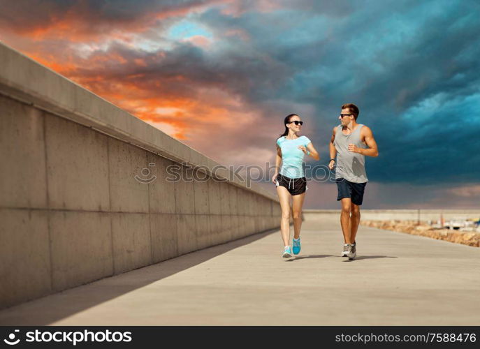 fitness, sport and people concept - happy couple in sports clothes and sunglasses running along pier over sunset sky on background. couple in sports clothes running along pier