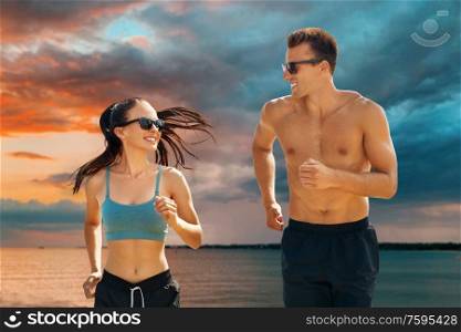 fitness, sport and people concept - happy couple in sports clothes and sunglasses running over sea and sunset sky on background. couple in sports clothes running along over sea