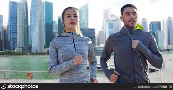 fitness, sport and people concept - happy couple running and listening to music in earphones over singapore city marina background. couple with earphones running over singapore 