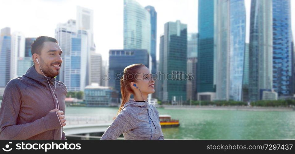fitness, sport and people concept - happy couple running and listening to music in earphones over singapore city marina background. happy couple with earphones running over singapore