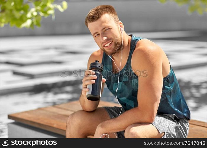 fitness, sport and people concept - happy smiling young man with earphones and bottle of water sitting on city bench. sportsman with earphones and bottle in city