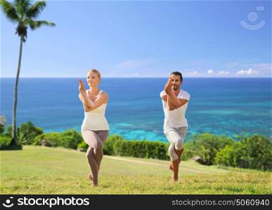 fitness, sport and people concept - smiling couple making yoga in eagle pose outdoors over exotic natural background with palm tree and ocean. smiling couple making yoga exercises outdoors