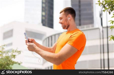 fitness, sport and people concept - smiling young man with smartphone and earphones listening to music at summer over city street on background. smiling young man with smartphone and earphones