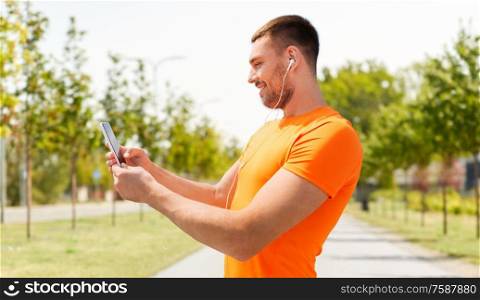 fitness, sport and people concept - smiling young man with smartphone and earphones listening to music at summer over city street on background. smiling young man with smartphone and earphones