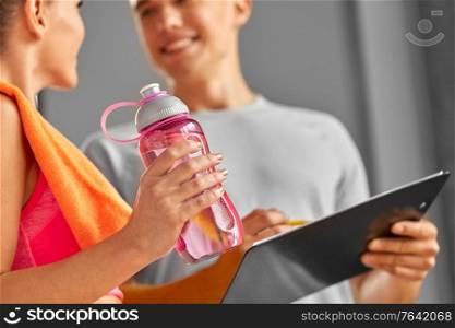 fitness, sport and people concept - smiling young woman with bottle of water and personal trainer with clipboard in gym. woman with water bottle and trainer in gym