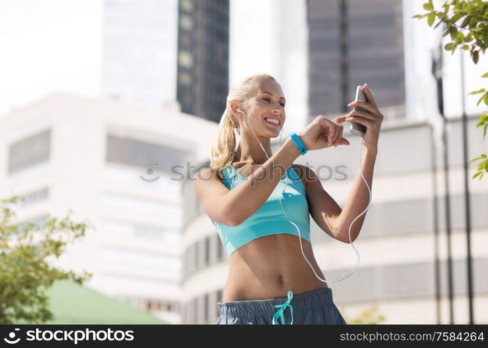 fitness, sport and people concept - smiling young woman with smartphone and earphones listening to music and exercising over city street on background. happy woman with smartphone and earphones at city