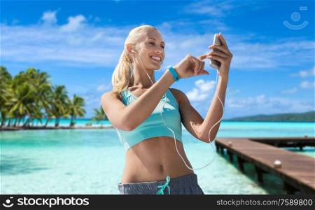 fitness, sport and people concept - smiling young woman with smartphone and earphones listening to music and exercising over wooden pier, palm trees and sea in french polynesia on background. happy woman with smartphone and earphones on beach