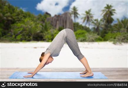 fitness, sport and people concept - woman doing yoga in downward facing dog pose on mat over exotic tropical beach background. woman doing yoga dog pose on beach