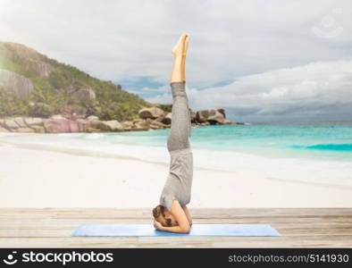 fitness, sport and people concept - woman doing yoga in headstand pose on mat over exotic tropical beach background. woman doing yoga in headstand pose on beach