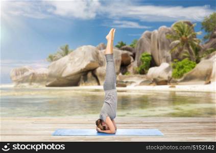 fitness, sport and people concept - woman doing yoga in headstand pose on mat over exotic tropical beach background. woman doing yoga in headstand pose on beach