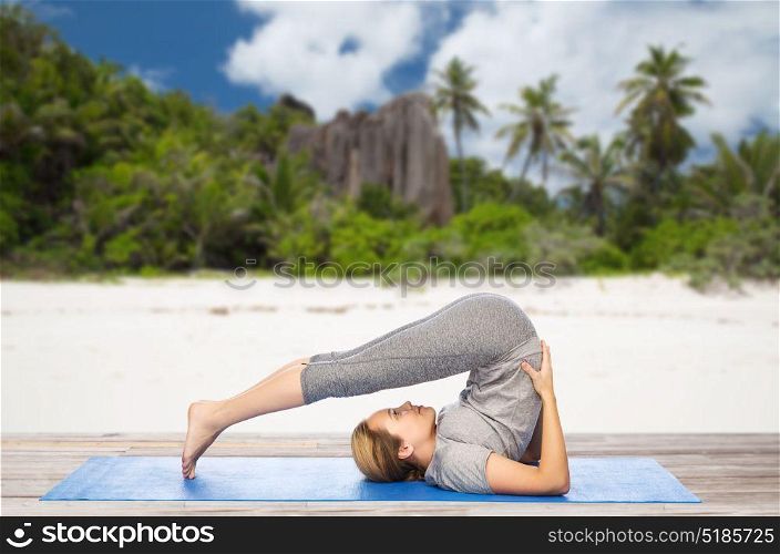 fitness, sport and people concept - woman doing yoga in plow pose on mat over exotic tropical beach background. woman doing yoga in plow pose on beach