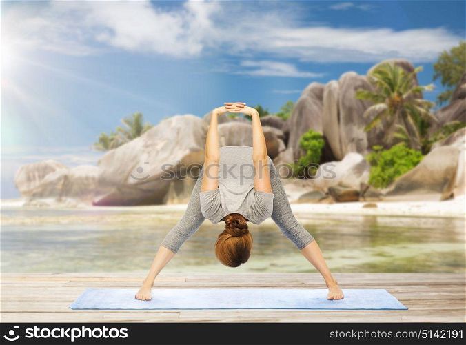 fitness, sport and people concept - woman doing yoga in wide-legged forward bend pose on mat over exotic tropical beach background. woman doing yoga wide-legged forward bend on beach