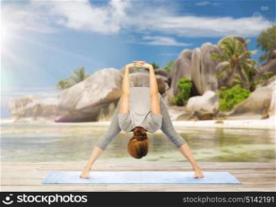 fitness, sport and people concept - woman doing yoga in wide-legged forward bend pose on mat over exotic tropical beach background. woman doing yoga wide-legged forward bend on beach