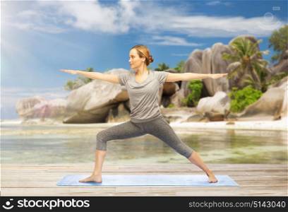 fitness, sport and people concept - woman doing yoga warrior pose on mat over exotic tropical beach background. woman doing yoga warrior pose on beach