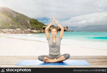 fitness, sport and people concept - woman meditating in yoga lotus pose on mat over exotic tropical beach background. woman meditating in yoga lotus pose on beach