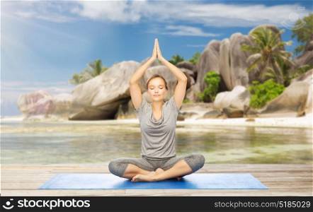 fitness, sport and people concept - woman meditating in yoga lotus pose on mat over exotic tropical beach background. woman meditating in yoga lotus pose on beach