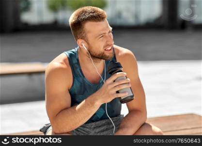 fitness, sport and people concept - young man with earphones and bottle of water sitting on city bench. sportsman with earphones and bottle in city