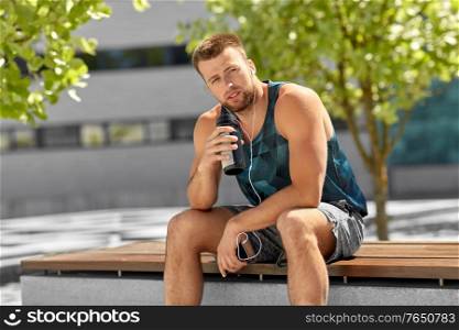 fitness, sport and people concept - young man with earphones, smartphone and bottle of water sitting on city bench. sportsman with earphones and bottle in city