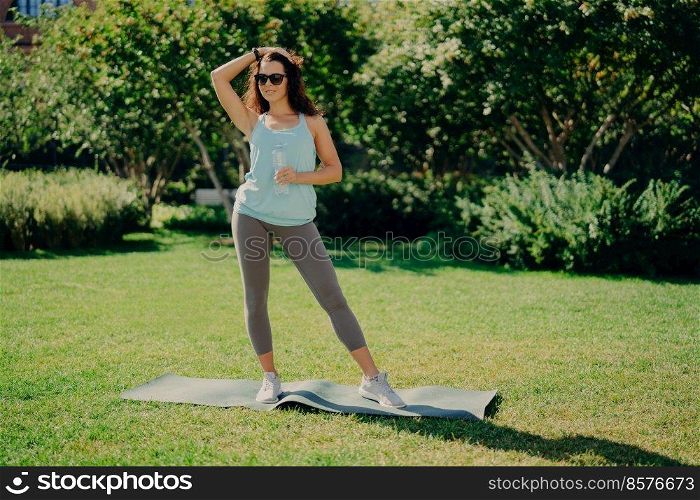 Fitness sport and recreation concept. Full length shot of sporty woman dressed in t shirt leggings and sneakers stands on karemat holds bottle of water poses against green nature background.