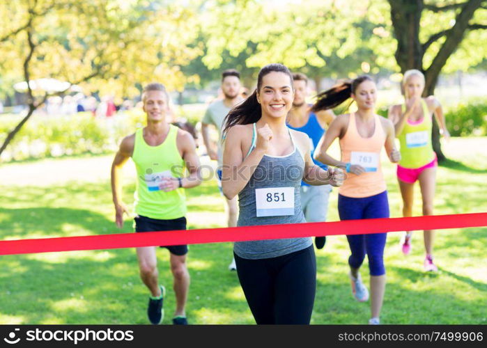 fitness, sport and success concept - happy teenage girl winning race and coming first to finish red ribbon over group of sportsmen running marathon with badge numbers at park. happy young female runner on finish winning race
