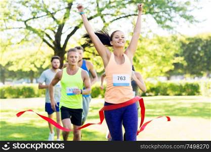 fitness, sport and success concept - happy woman winning race and coming first to finish red ribbon over group of sportsmen running marathon with badge numbers at park. happy young female runner on finish winning race