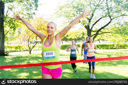fitness, sport and success concept - happy woman winning race and coming first to finish red ribbon over group of sportsmen running marathon with badge numbers at park. happy young female runner on finish winning race