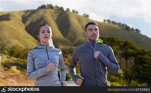 fitness, sport and technology concept - happy couple running and listening to music in earphones over big sur hills background in california. couple with earphones running over big sur. couple with earphones running over big sur