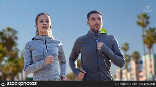 fitness, sport and technology concept - happy couple running and listening to music in earphones over venice beach background in california. couple with earphones running over venice beach. couple with earphones running over venice beach